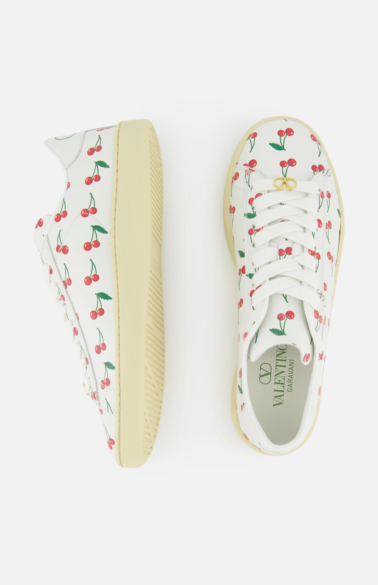 The VALENTINO GARAVANI Royco Sneakers feature white leather, cream-colored soles, and a playful red cherry pattern with green leaves. Shown from the side and top on a white background.