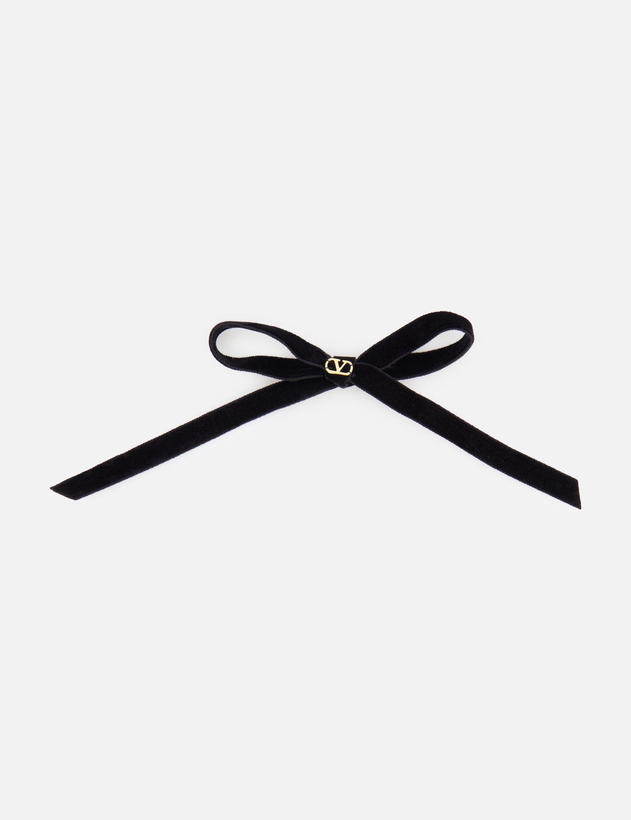 The VALENTINO GARAVANI Vlogo Signature Hairclip Small Bow is a black velvet hair clip with a simple bow and a gold “V” emblem at the center, shown on a white background.