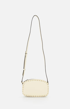 The VALENTINO GARAVANI Rockstud Camera Case Bag is a cream oval handbag with gold-tone studs along the edges and an adjustable long strap, shown against a plain white background.
