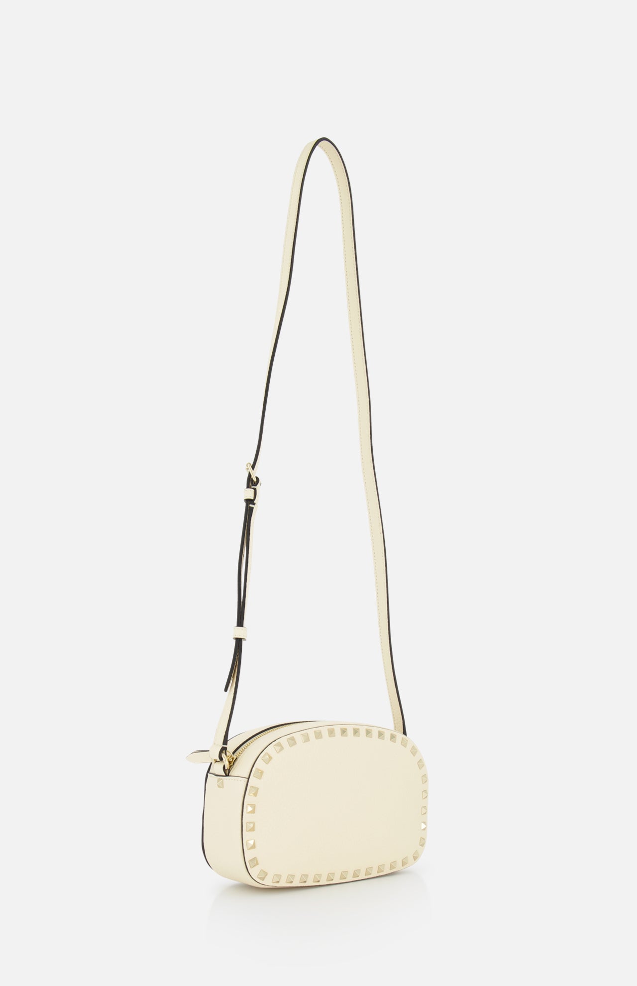 The VALENTINO GARAVANI Rockstud Camera Case Bag is a small, oval ivory leather crossbody with a long adjustable strap and gold stud accents around the edges, shown on a white background.
