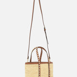 The VALENTINO GARAVANI Rockstud Mini Tote Bag features woven raffia, brown leather top handles, an adjustable shoulder strap, and gold-tone rockstud accents. It's shown against a plain white background.