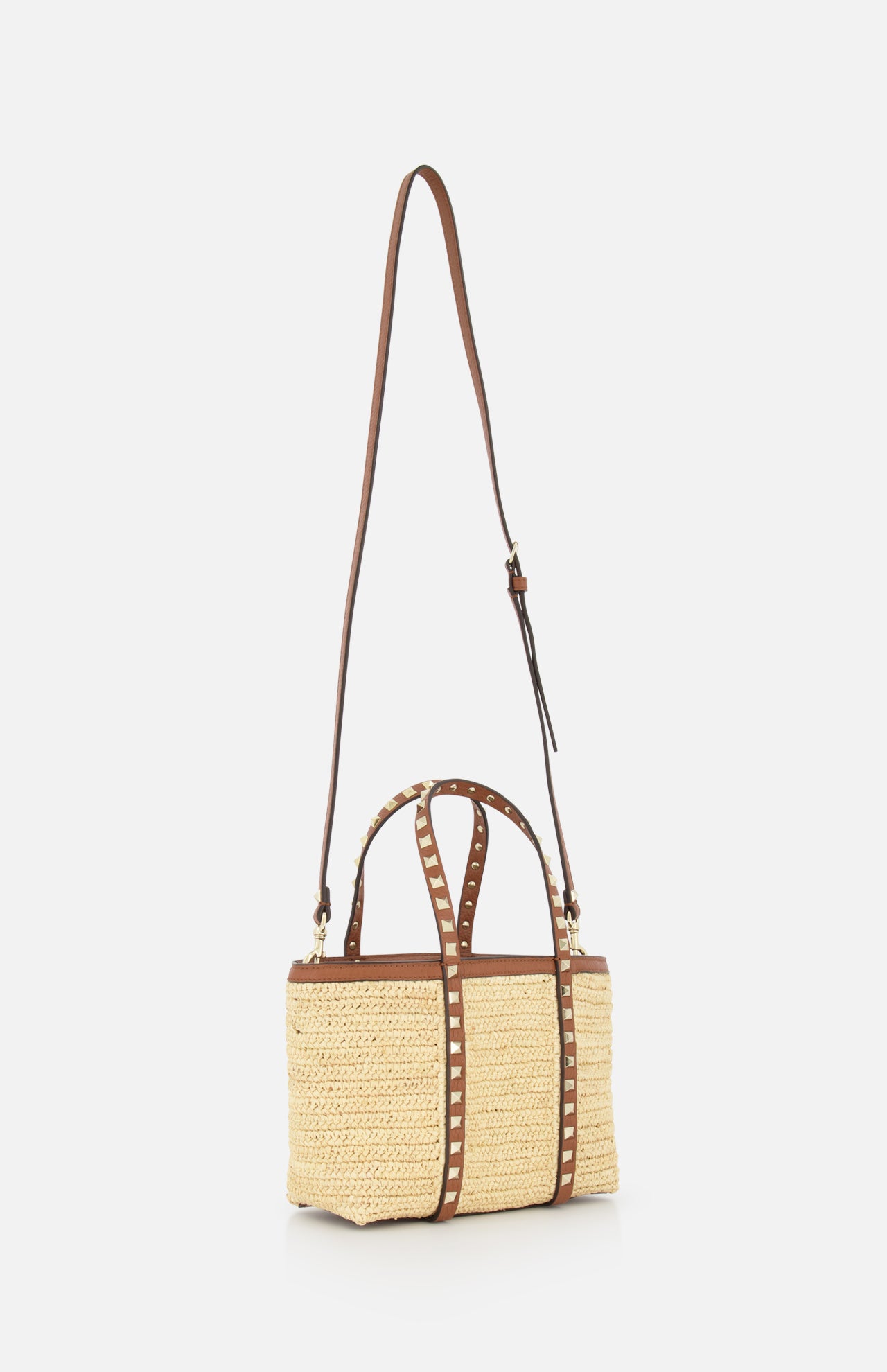 The VALENTINO GARAVANI Rockstud Mini Tote Bag features woven raffia, brown leather top handles, an adjustable shoulder strap, and gold-tone rockstud accents. It's shown against a plain white background.