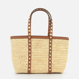 The VALENTINO GARAVANI Rockstud Mini Tote Bag is crafted from woven raffia and leather with brown handles, gold rockstud accents, and an open-top rectangular design for easy access.