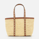 The VALENTINO GARAVANI Rockstud Mini Tote Bag features woven raffia and leather with brown handles and gold rockstud accents. This rectangular, open-top bag is shown against a plain white background.