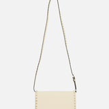 The VALENTINO GARAVANI Rockstud Small Shoulder Bag, crafted in cream-colored leather with a long adjustable strap and small square stud details along the edges, is elegantly displayed on a white background.