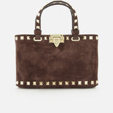 The Rockstud Suede Mini Bag by VALENTINO GARAVANI is a rectangular, dark chocolate brown suede mini bag, embellished with gold pyramid studs and a matching gold clasp. It features two handles accented with gold studs and includes a removable chain strap, all elegantly displayed on a white background.