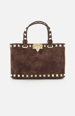 The Rockstud Suede Mini Bag by VALENTINO GARAVANI is a rectangular, dark chocolate brown suede mini bag, embellished with gold pyramid studs and a matching gold clasp. It features two handles accented with gold studs and includes a removable chain strap, all elegantly displayed on a white background.
