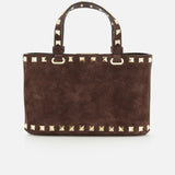 The VALENTINO GARAVANI Rockstud Suede Mini Bag is crafted in a rich dark chocolate brown suede, featuring two handles and distinctive gold square studs accenting the edges and handles. Displayed against a white background, this mini bag also comes with a removable chain strap for versatile styling options.
