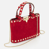 The VALENTINO GARAVANI Rockstud Suede Mini Bag features red suede, gold studded edges, a matching handle, gold clasp, and a detachable chain strap, shown against a white background.
