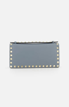 The VALENTINO GARAVANI Rockstud Wallet with Chain Strap is a rectangular, light blue leather crossbody bag featuring gold pyramid studs along the edges, perfect as a chic party purse and displayed against a plain white background.