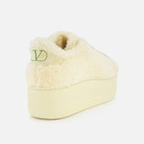 The VALENTINO GARAVANI Royco Shearling Platform Sneaker is shown from the back, highlighting its cream-colored shearling construction and thick platform heel. A green V-shaped logo detail appears on the left side of the heel.