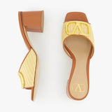 The VALENTINO GARAVANI V Logo Signature Block Heel Slide Sandal features tan and yellow woven raffia uppers, block heels, and gold decorative buckles, shown on a white background.