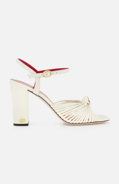 The Valentino Garavani Annet T.90 Sandal by VALENTINO GARAVANI features white knotted leather straps, a leather ankle strap with gold buckle, an ivory high heel, and a subtle VLogo accent near the sole.