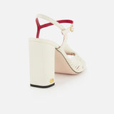 The Valentino Garavani Annet T.90 Sandal features a leather ankle strap with gold buckle, ivory block heel, red-trimmed back strap, and a signature VLogo near the heel base by VALENTINO GARAVANI.