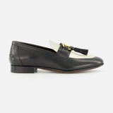 The Valentino Garavani Vaniteux Loafer by VALENTINO GARAVANI is a black and white women's leather loafer with a low heel, gold metal bit, and black tassel detail, offering classic slip-on elegance against a plain white background.