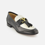 The Valentino Garavani Vaniteux Loafer by VALENTINO GARAVANI features black and white leather, a gold decorative emblem, and black tassels on the front, shown here against a white background.