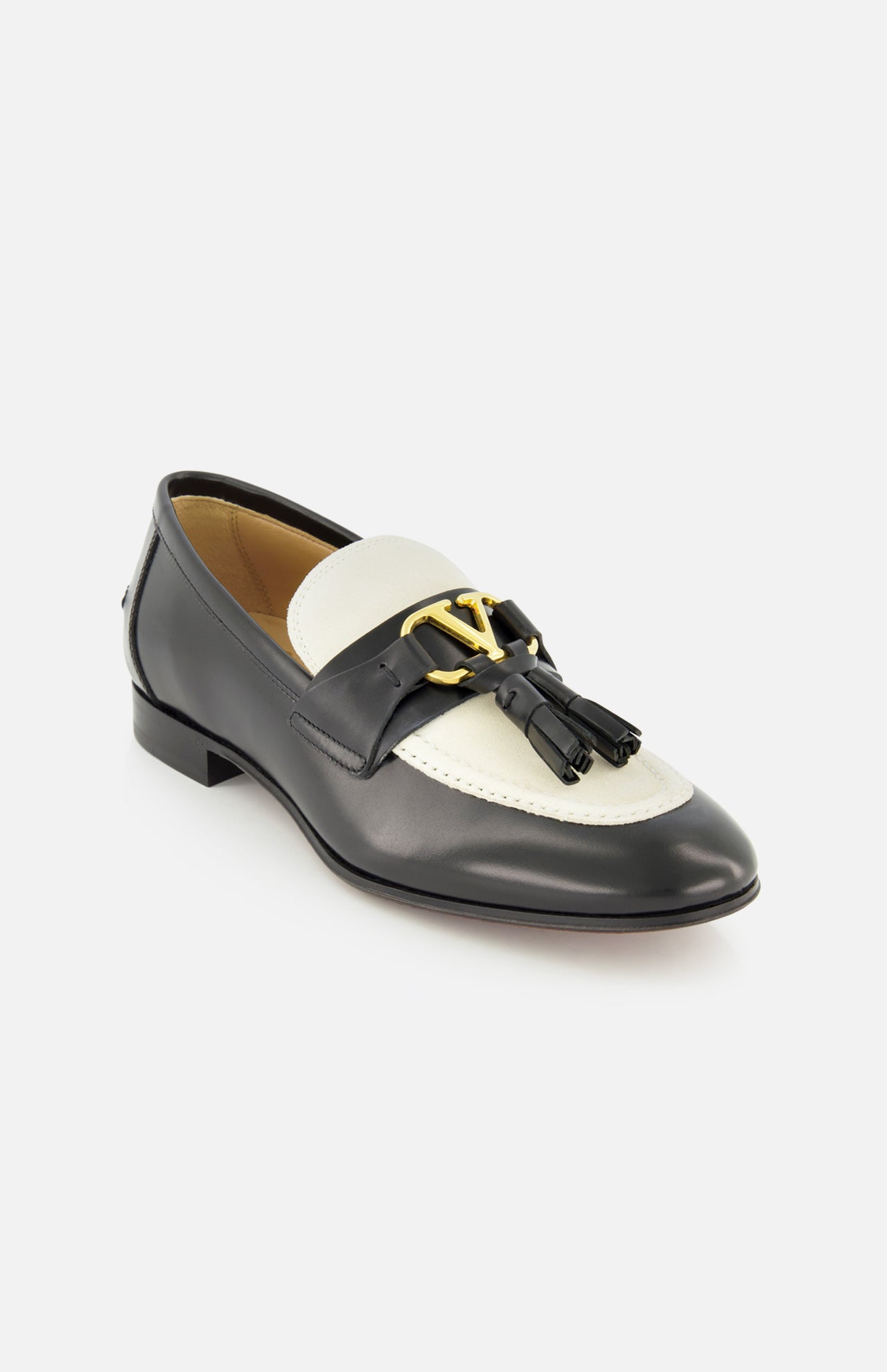 The Valentino Garavani Vaniteux Loafer by VALENTINO GARAVANI features black and white leather, a gold decorative emblem, and black tassels on the front, shown here against a white background.
