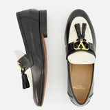 The VALENTINO GARAVANI Vaniteux Loafer is a women's black and white leather slip-on with tassels and gold hardware, featuring classic style and modern elegance, shown from side and top views on a white background.