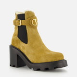 The VLogo Signature Beatle Boot by VALENTINO GARAVANI features tan suede with a chunky black heel and sole, gold buckle detail, black elastic side panel, and visible shearling lining at the top—these chic ankle boots offer a stylish twist on classic design.