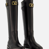 The VALENTINO GARAVANI VLogo Signature Boot is a pair of knee-high black leather boots with low heels and gold “V” logo buckles near the cuffs, perfect for adding equestrian style to any outfit. Shown against a plain white background.