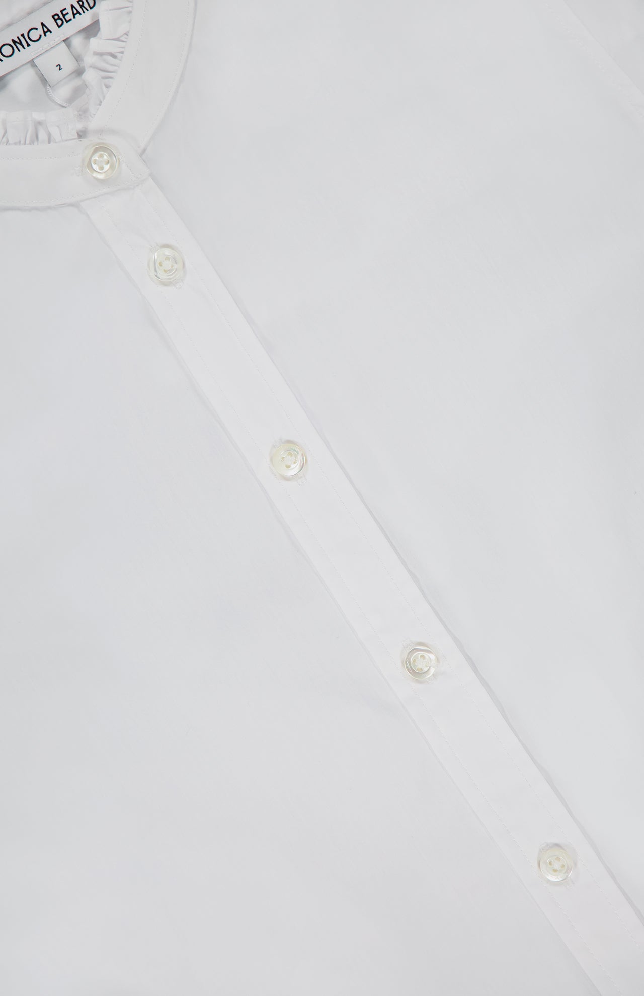 Close-up of the VERONICA BEARD Bays Ruffle Collar Shirt, a crisp white button-down with matching buttons and a visible collar label. Made from stretch cotton, this shirt offers a smooth finish that's ideal for feminine dressing.