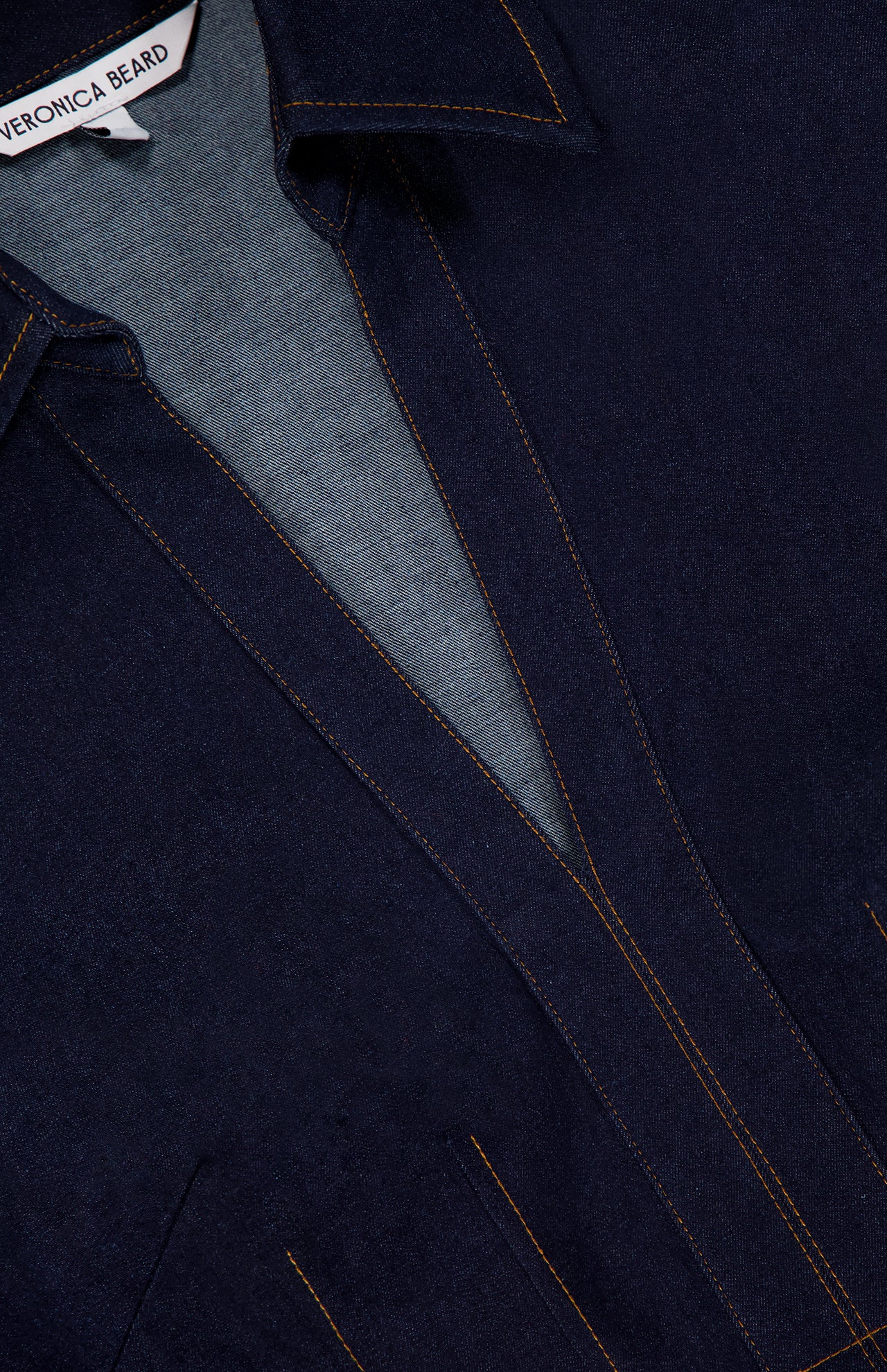 Close-up of the Raymond Denim Mini Dress by VERONICA BEARD, featuring dark blue denim, orange stitching, a fit-and-flare silhouette, and a partially visible brand label at the neckline.