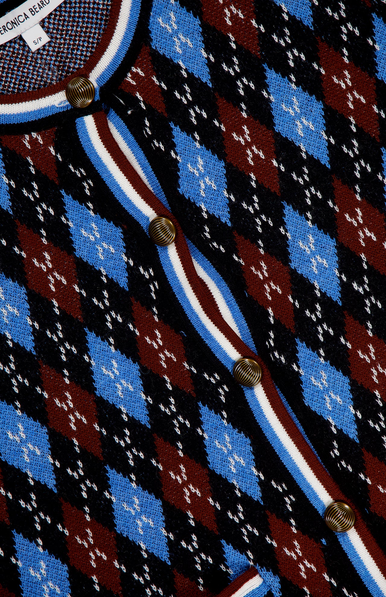 A close-up of the VERONICA BEARD Sylvane Cropped Argyle Cardigan, showcasing a slim fit, blue, black, and burgundy diamond pattern, gold round buttons, and a white, burgundy, and navy striped trim with the label near the collar.