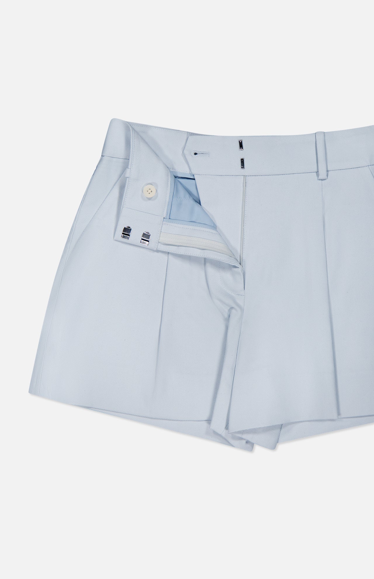 The Craig High Rise Short by VERONICA BEARD in light blue Italian cotton-blend twill features pleats, belt loops, a white inner waistband button, metal hooks, and zip closure, shown partially unbuttoned and unzipped.