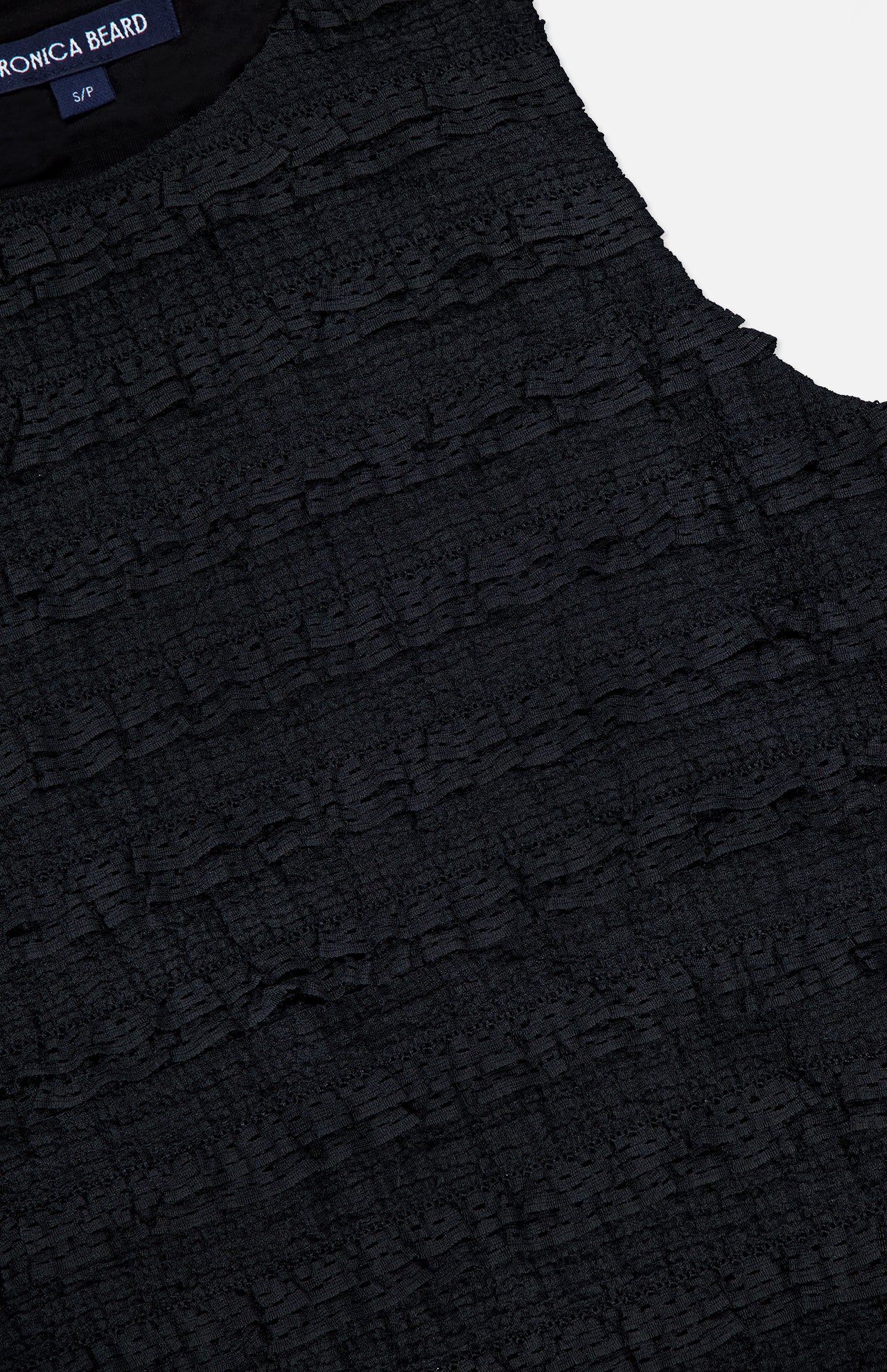 Close-up of the VERONICA BEARD Ruffled Cropped Jordyn Tank, a sleeveless black top crafted from textured stretch jersey with horizontal micro-ruffles. A navy blue label with white text appears at the neckline.