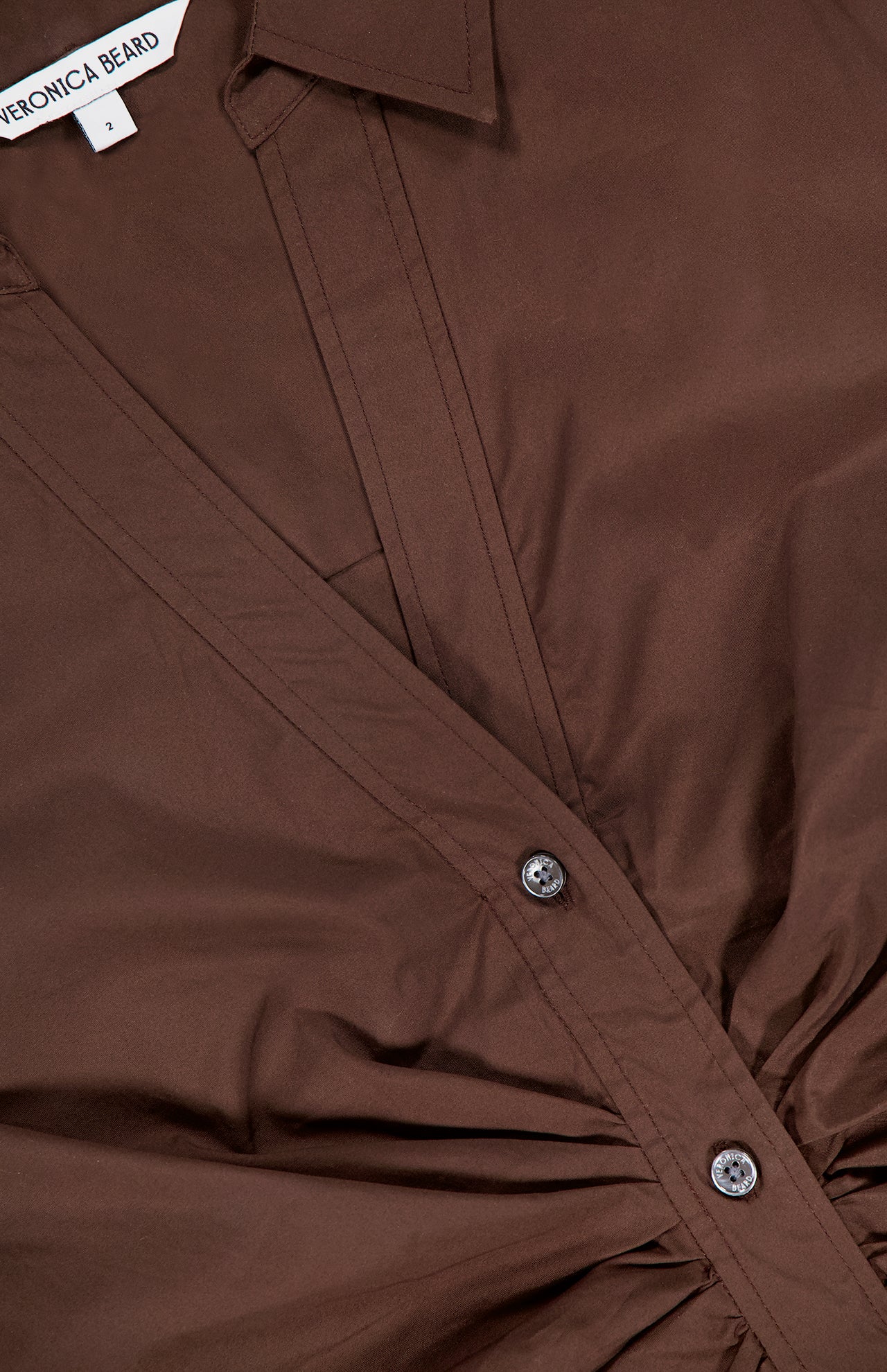 A close-up of the VERONICA BEARD Devra Short Sleeve Ruched Top in brown stretch cotton poplin with button details and waist-defining ruching, labeled VERONICA BEARD, size 2.