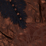 Close-up of the Pascaline Floral Lace Top by VERONICA BEARD, showcasing brown jacquard fabric with a floral motif, black lace trim, and brown decorative buttons arranged vertically.