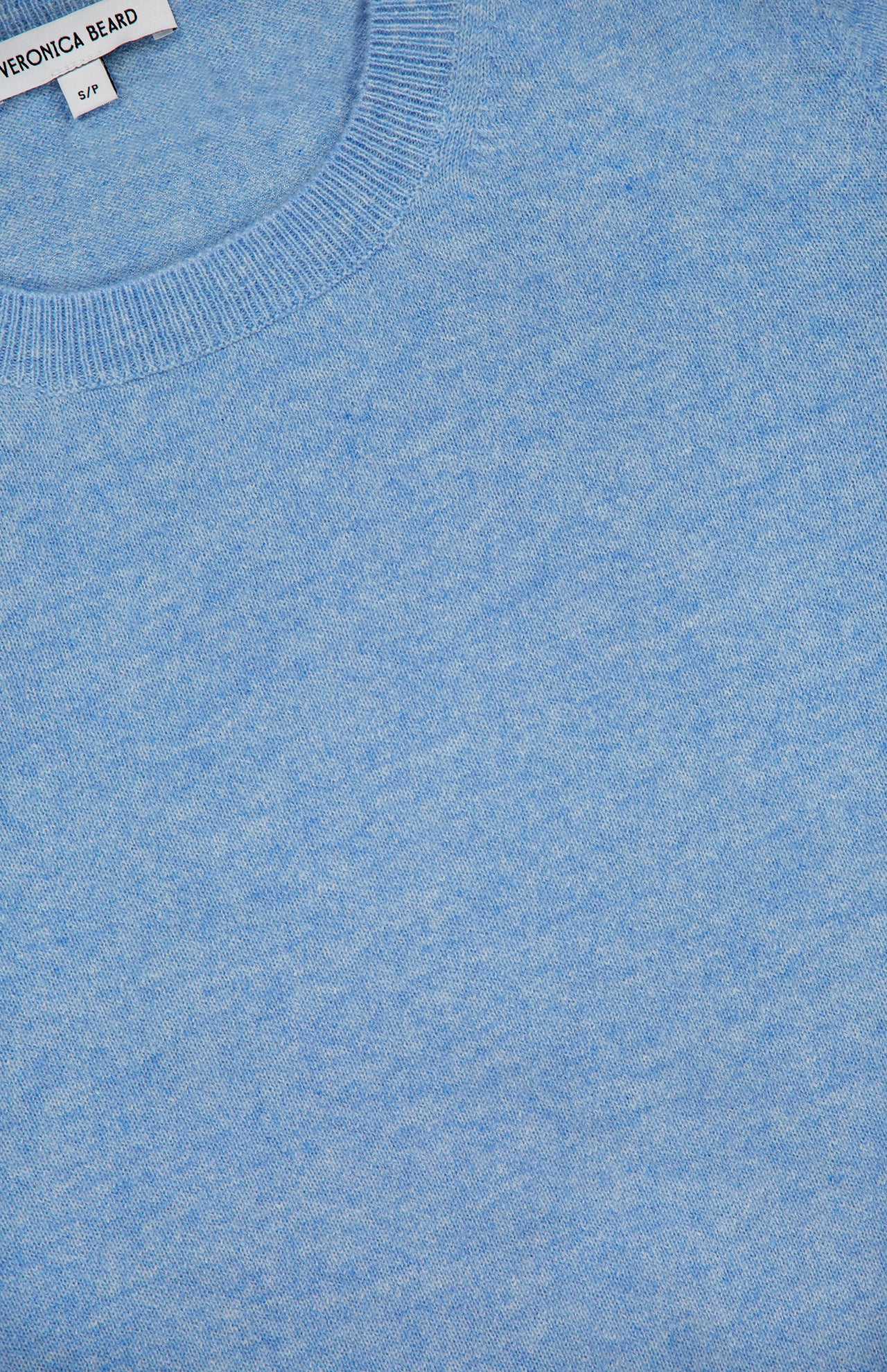 Close-up of the Shana Cropped Cashmere Sweater by VERONICA BEARD in light blue, highlighting its knit texture and neckline. A portion of the garment label with the brand name and size S/P appears at the top left corner. This relaxed fit sweater features a subtly cropped hem, offering a modern silhouette.