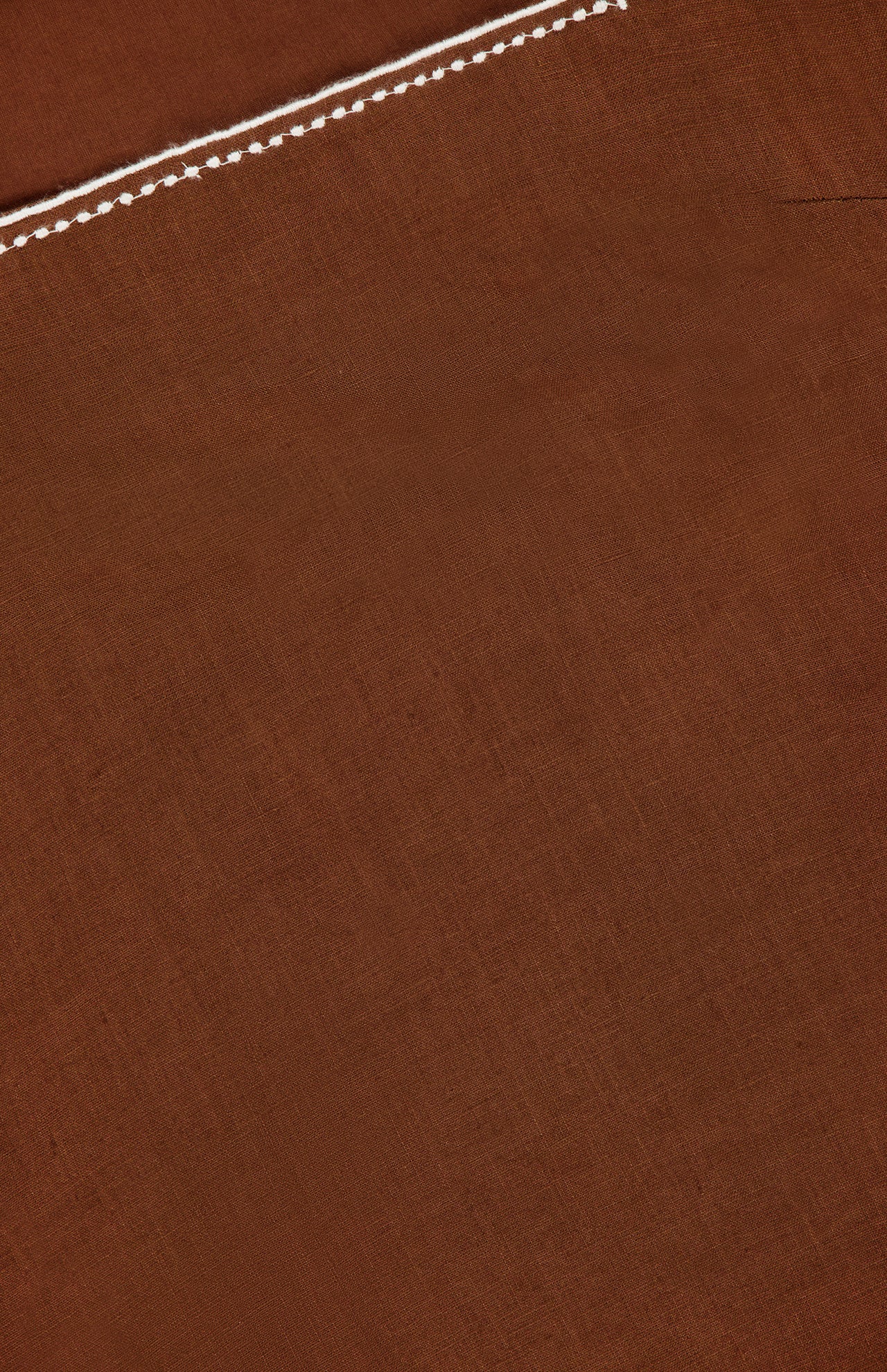 A piece of brown linen fabric features a stitched top seam, diagonal crease, and embroidered trim details inspired by the Shanice Embroidered Mini Dress by VERONICA BEARD.