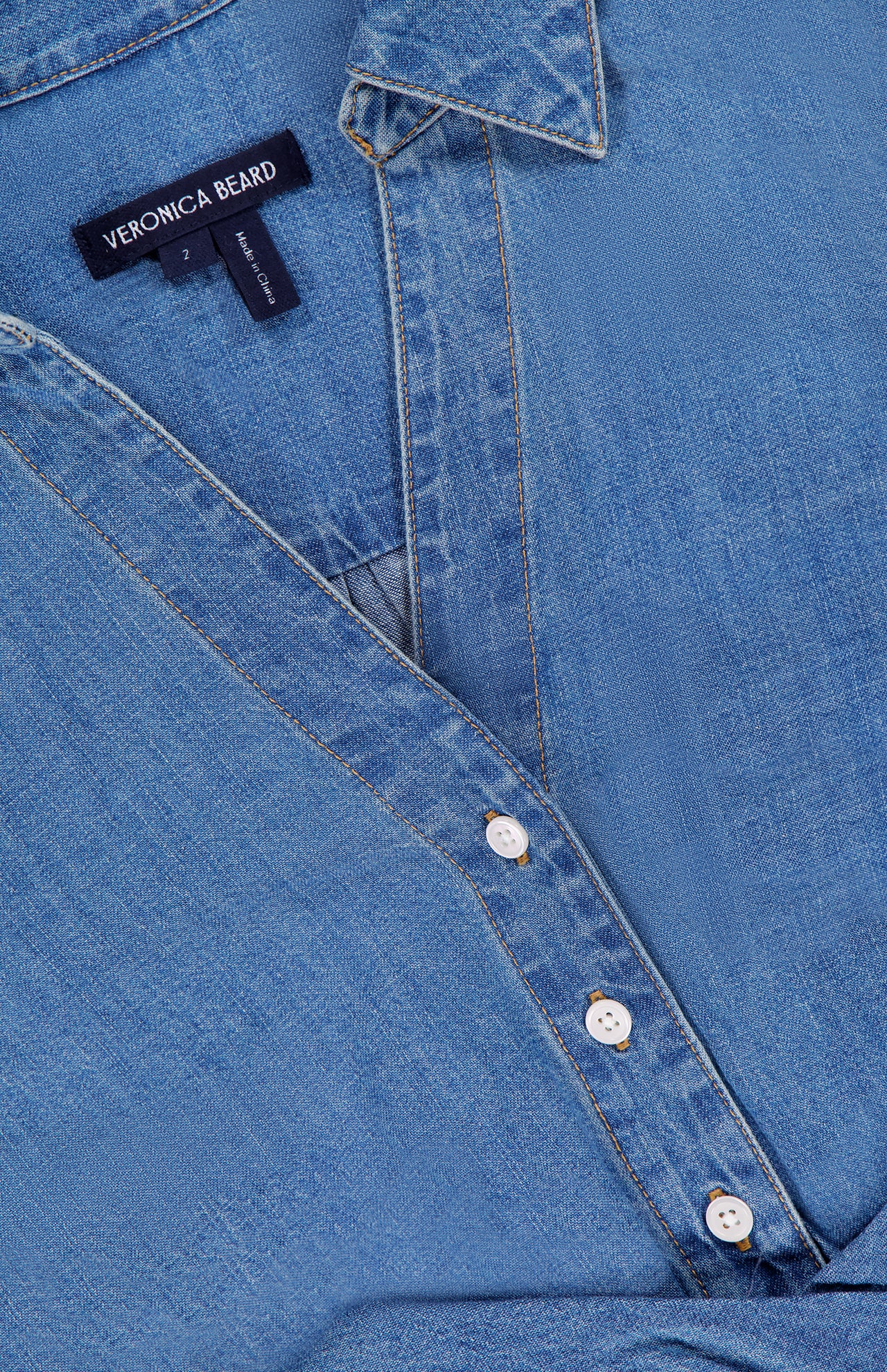 Close-up of the Timothy Twist Detail Shirt by VERONICA BEARD—a blue jean shirt with light brown stitching, white buttons, label inside the collar, and a subtle twist waist detail.