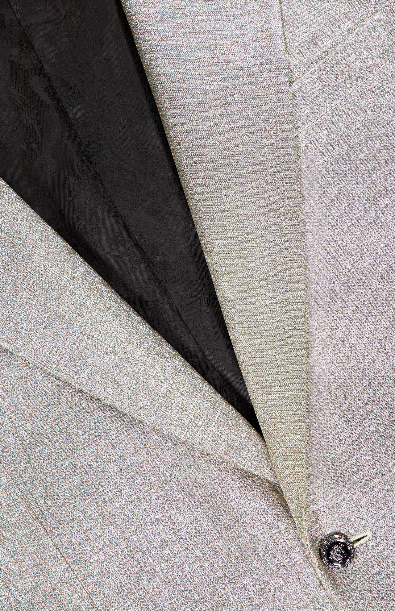 Close-up of the VERSACE Metallic Evening Tux Jacket, highlighting its light silver lurex texture, detailed lapel, black floral-patterned lining, buttonhole, and signature shiny Medusa button.