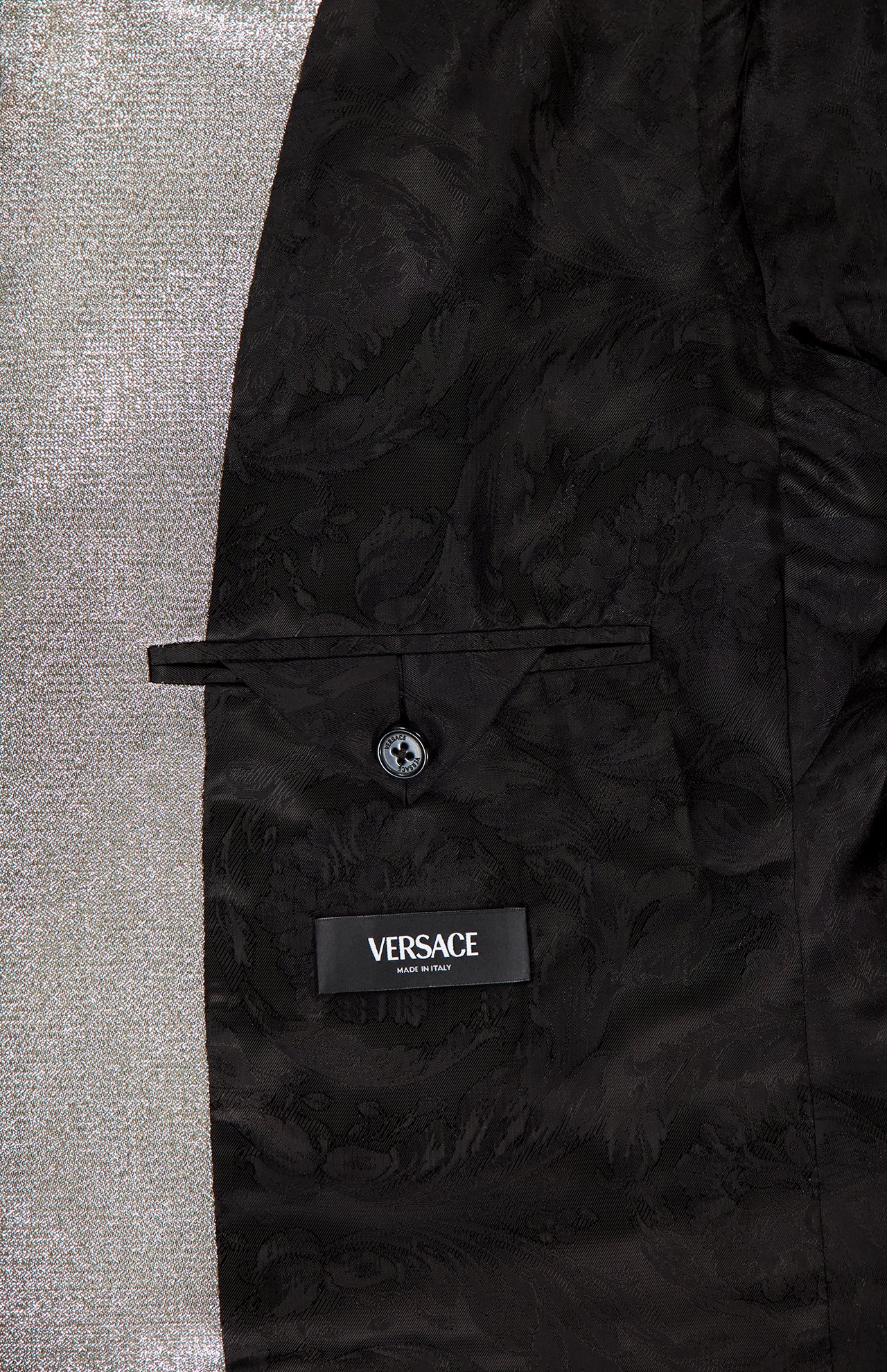 Close-up of the Metallic Evening Tux Jacket by VERSACE shows its elegant floral lining, buttoned pocket, and black VERSACE label. The luxurious interior contrasts with shiny silver fabric, emphasizing the jacket's sophisticated metallic style.