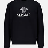 The Medusa Cashmere Knit Sweater by VERSACE is a black crewneck with long sleeves, showcasing the iconic Medusa logo and bold white VERSACE branding on the front—a standout in luxury Italian menswear.
