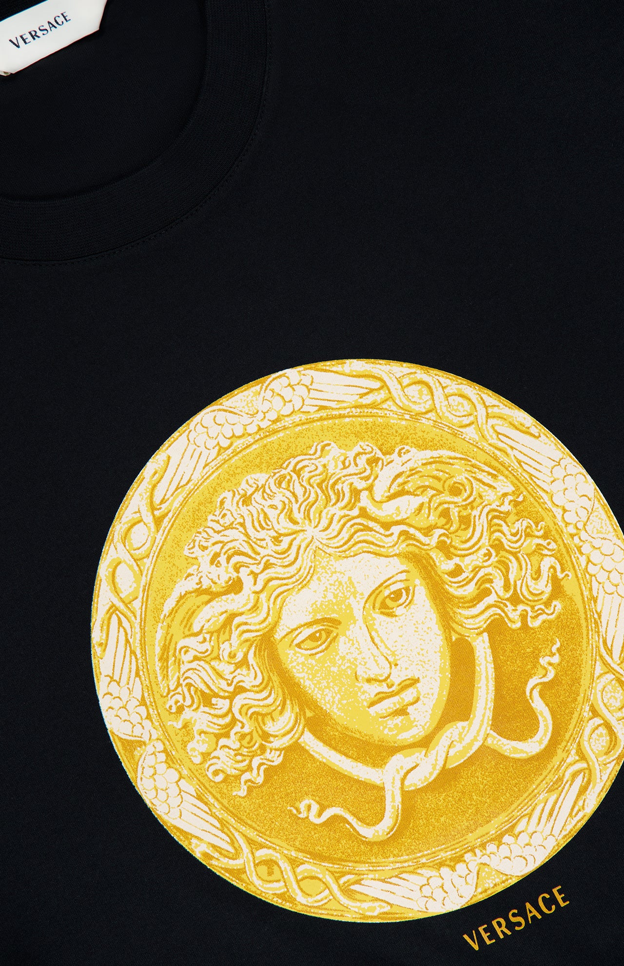 The Cotton Medusa Coin Jersey T-Shirt by VERSACE epitomizes luxury Italian men’s fashion with its bold central Medusa coin graphic, encircled by an ornate border. Featuring the iconic Versace logo in elegant gold lettering at the hem, this black t-shirt makes a striking addition to any contemporary wardrobe.
