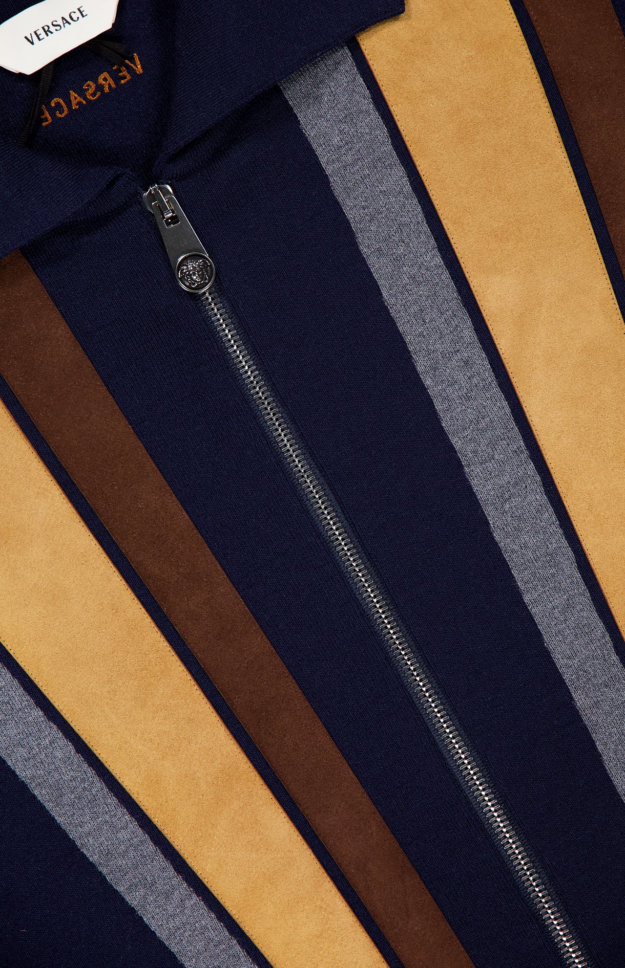Close-up of a VERSACE Leather Patchwork Knit Jacket with a vertical zipper and bold navy, tan, brown, and gray stripes. The VERSACE label at the top left underscores this luxury Italian menswear piece.