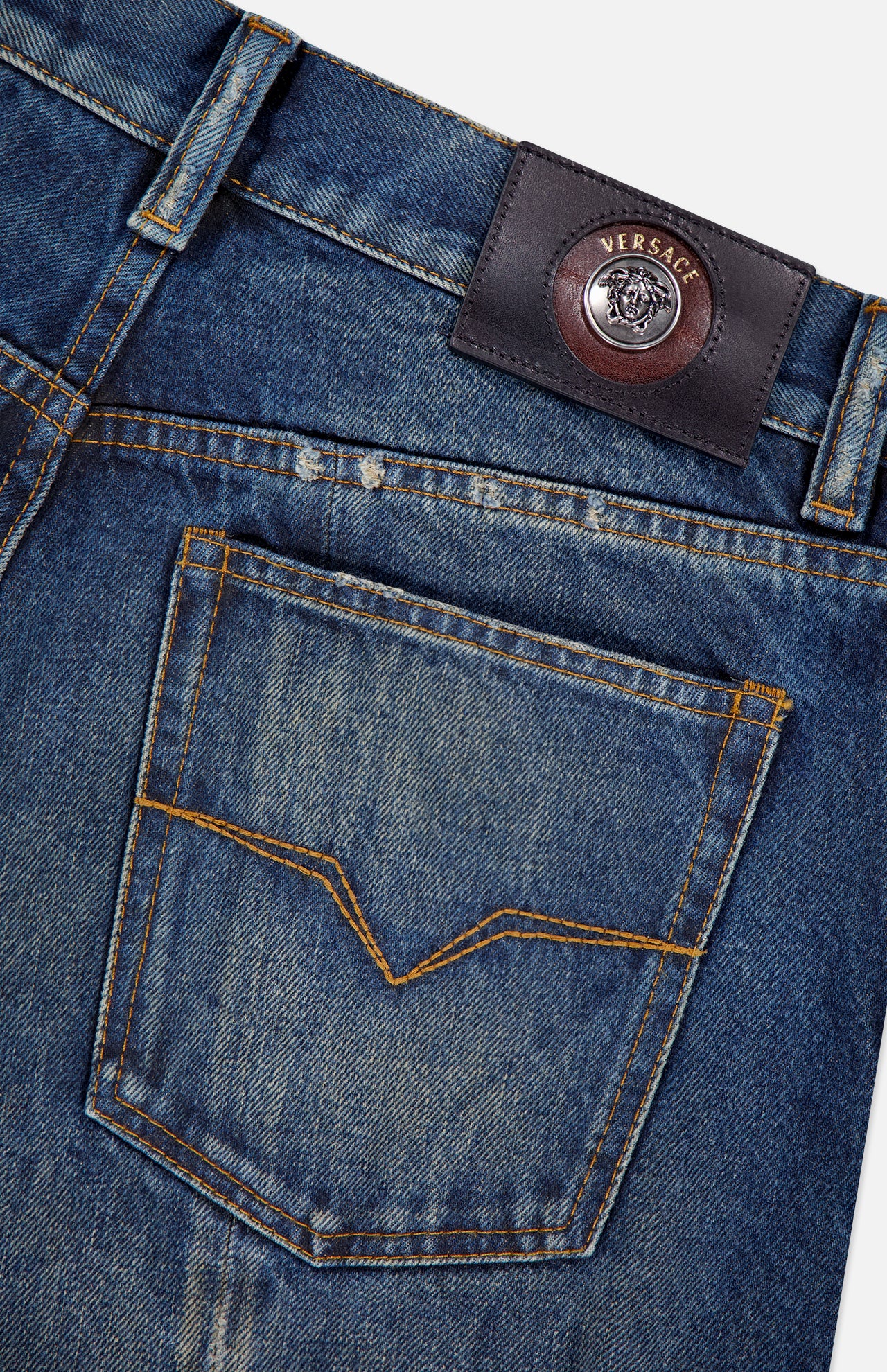 Close-up of VERSACE Non-Stretch Dirty Wash Jeans in Dirty Blue, showing a back pocket with yellow stitching and a black leather Medusa logo label above the waistband.