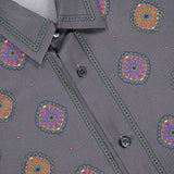 Close-up of the VERSACE Silk Twill Print The Suitor Shirt in gray silk, showcasing its vibrant red, blue, yellow, and green print with black buttons and detailed stitching along the collar and placket.