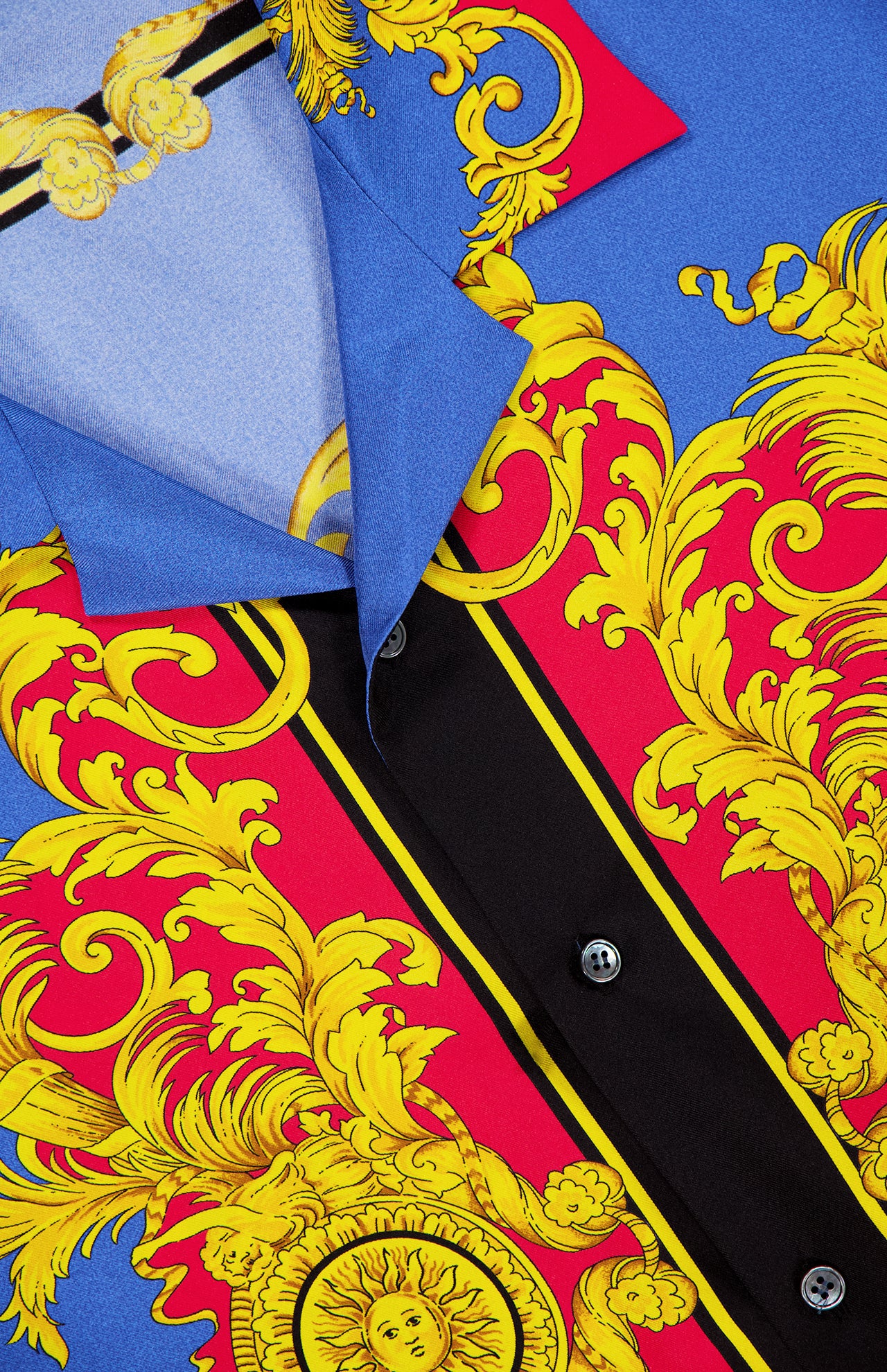 Close-up of VERSACE's Silk Twill Print The Sun Shirt, a luxury Italian menswear piece in blue and red silk with ornate yellow baroque print, black vertical stripes, sun motifs, black buttons, and a visible collar.
