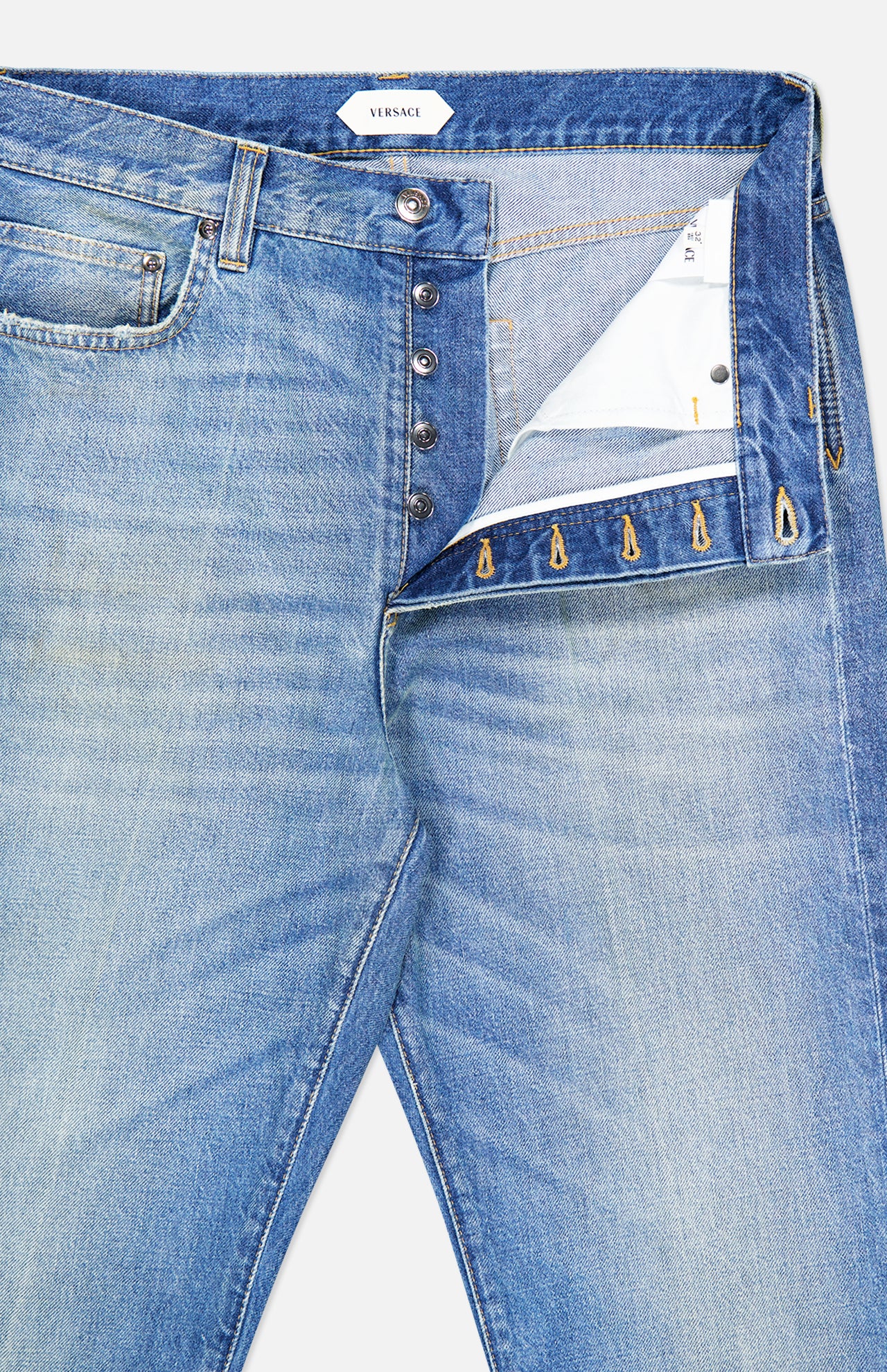 A close-up of the VERSACE Stone Washed Rigid Jeans with the fly open, featuring metallic buttons, a white inner lining, and a visible VERSACE label on the inner waistband.