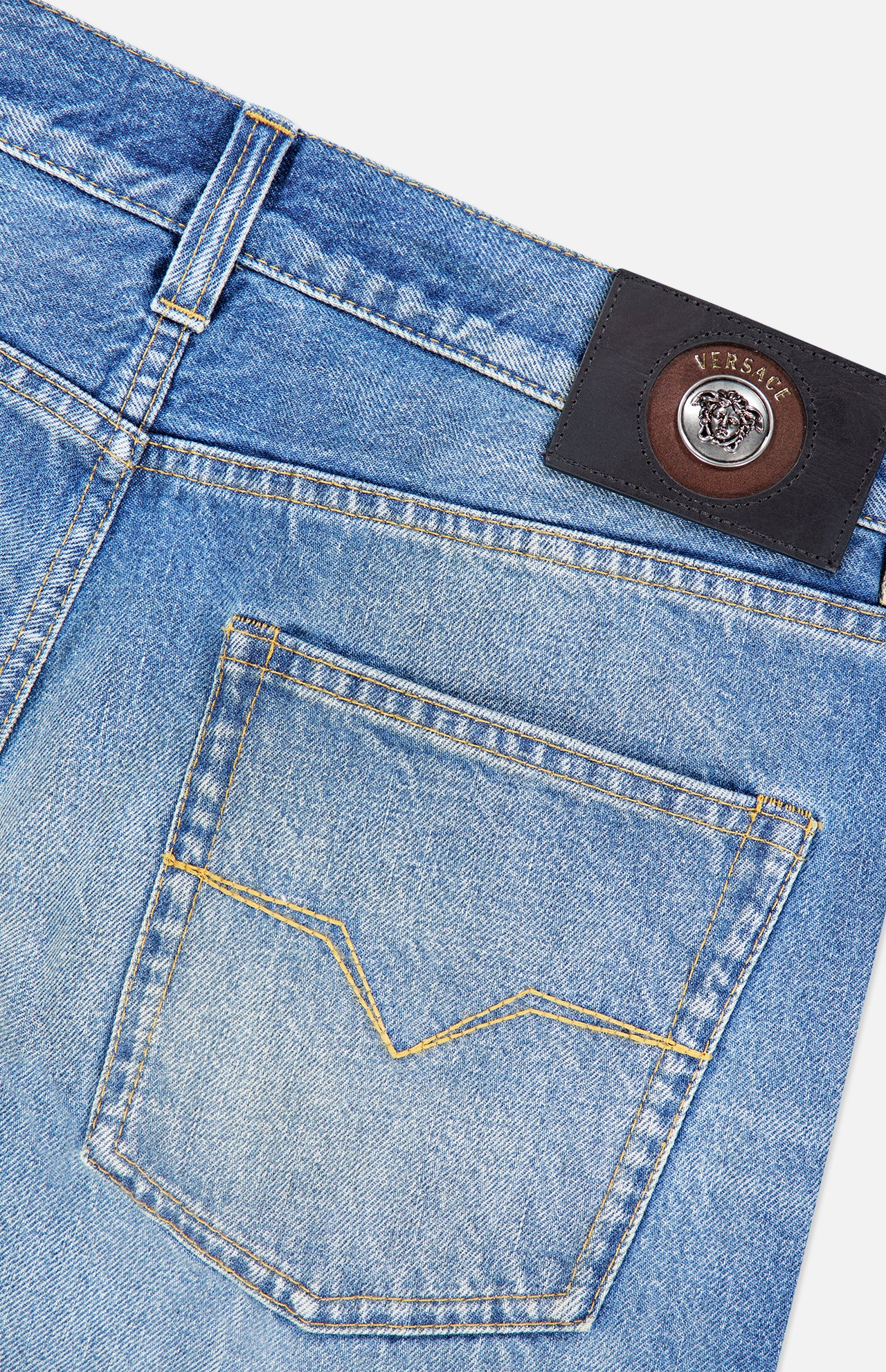 Close-up of light blue VERSACE Stone Washed Rigid Jeans featuring a back pocket with yellow stitching and a black leather VERSACE label with the Medusa logo on the waistband—an iconic detail of these men’s designer straight-leg jeans.