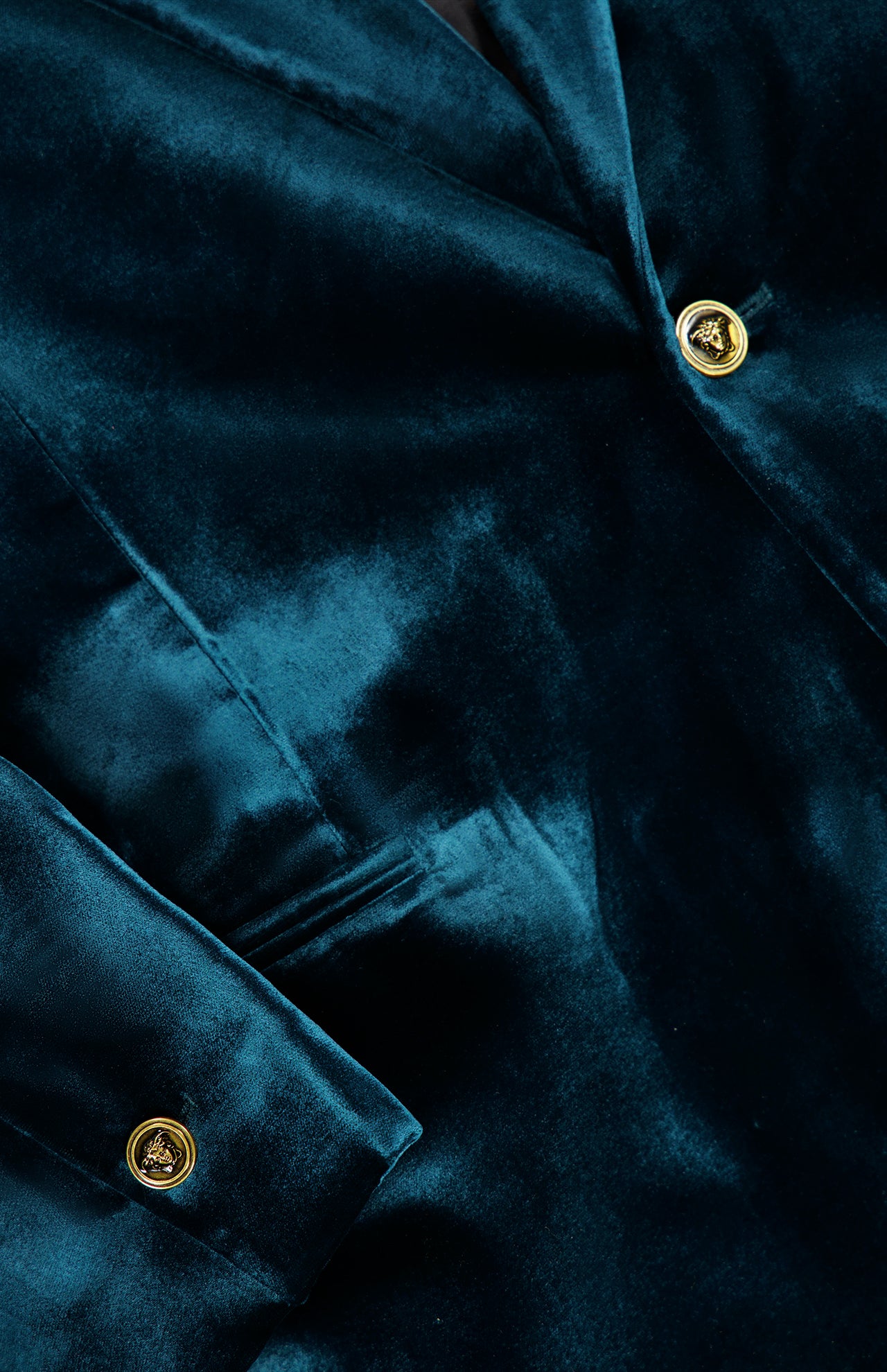 A close-up of the VERSACE Viscose Silk Liquid Velvet Evening Jacket showcases its men’s luxury design in deep green velvet, highlighted by gleaming gold buttons and a welt pocket that emphasize the sumptuous texture and rich, vibrant color of the fabric.