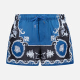 VERSACE Mythic Barocco Print Swim Shorts: blue and black with a white ornate floral and scroll baroque motif, featuring a drawstring waistband and mid-thigh length. Luxury Italian swimwear style.