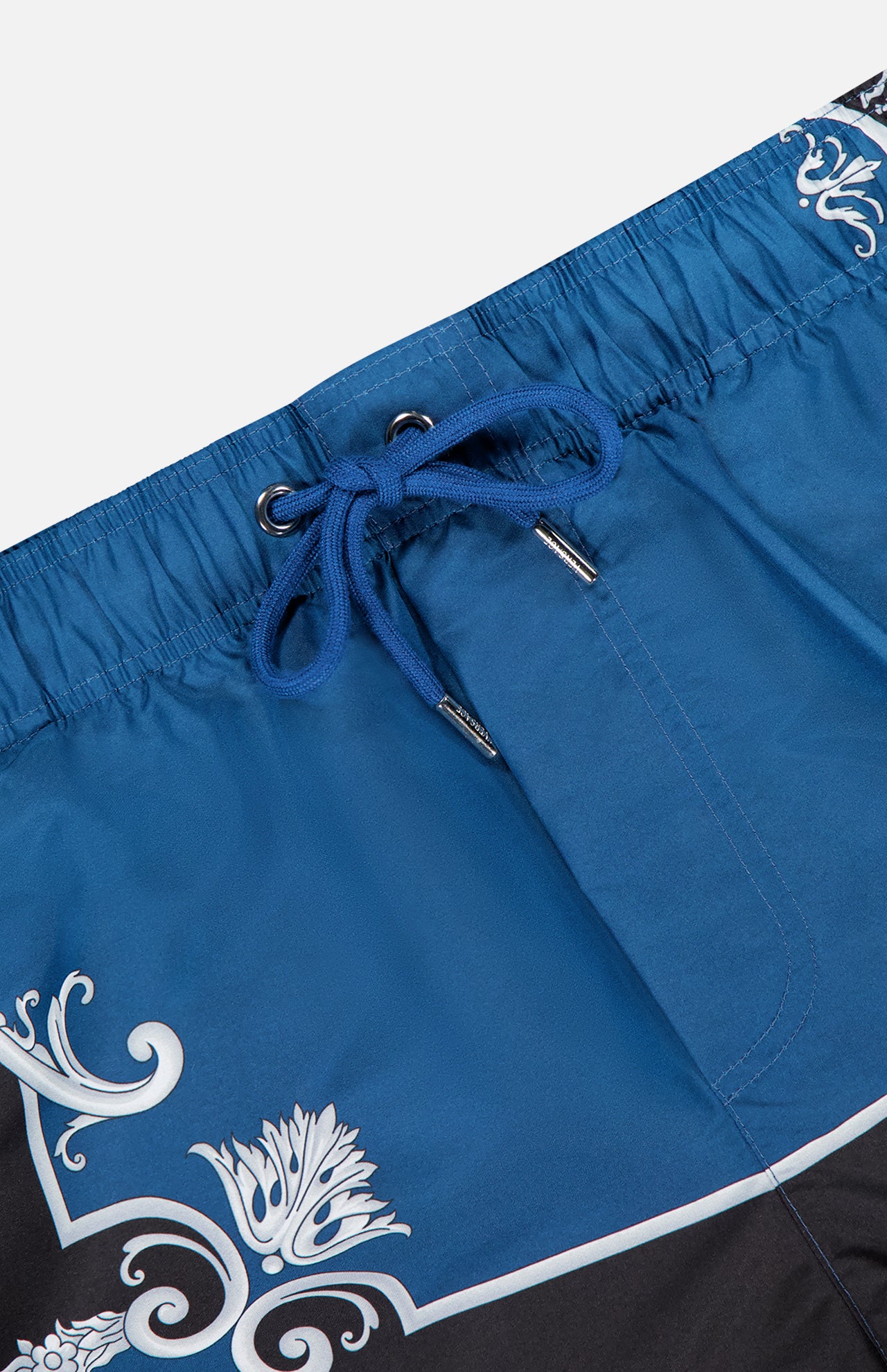 Close-up of VERSACE Mythic Barocco Print Swim Shorts in blue, featuring a white floral motif, silver eyelets, and fine stitching details that highlight the sophisticated Italian-inspired design.