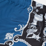 The VERSACE Mythic Barocco Print Swim Shorts feature blue and black fabric with ornate white baroque patterns, including a Medusa head and stylized flourishes—a classic luxury Italian swimwear design.