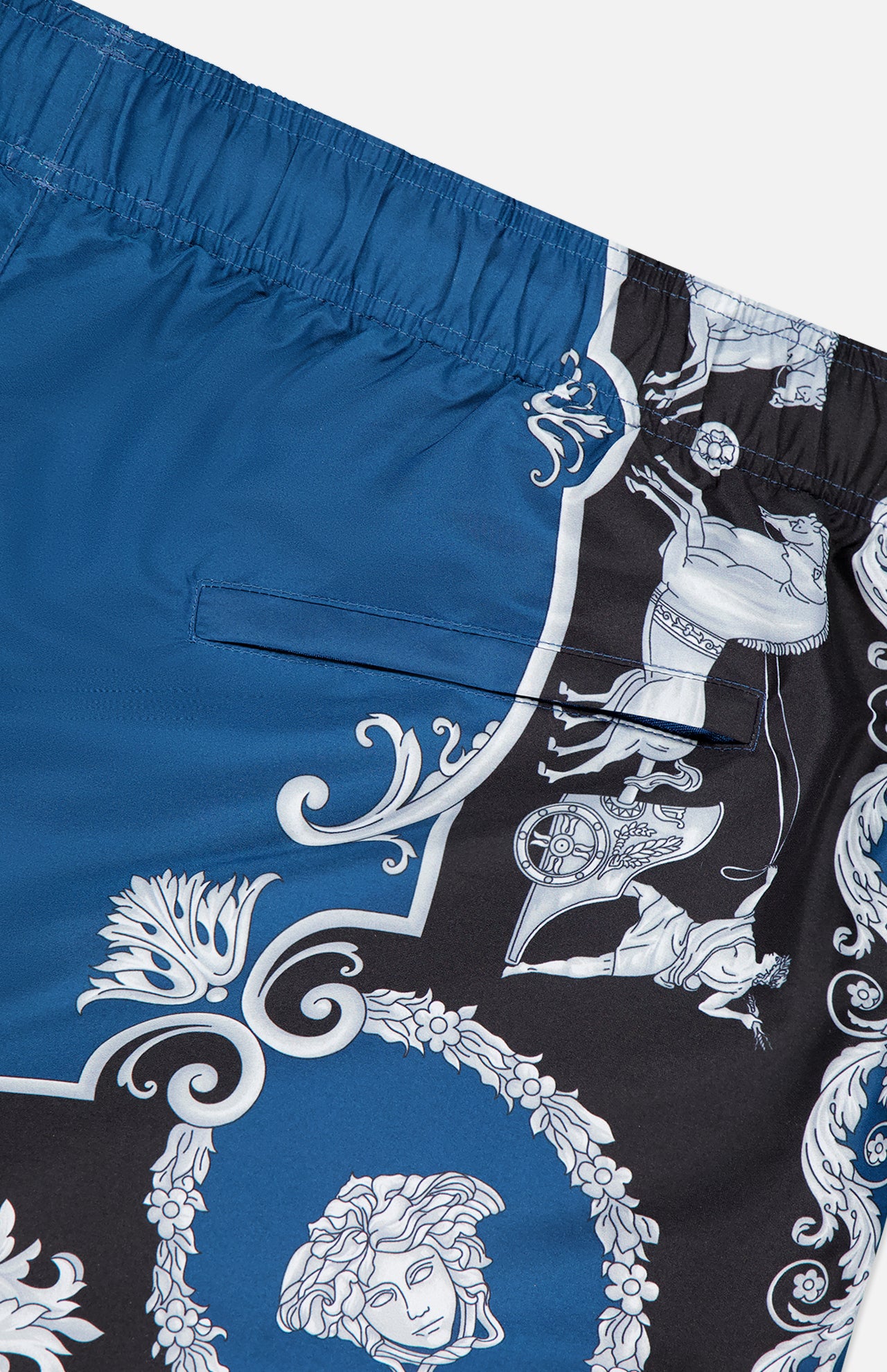 The VERSACE Mythic Barocco Print Swim Shorts feature blue and black fabric with ornate white baroque patterns, including a Medusa head and stylized flourishes—a classic luxury Italian swimwear design.