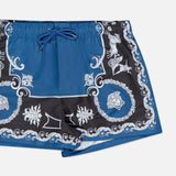The VERSACE Mythic Barocco Print Swim Shorts feature a blue and black baroque design with white ornate floral and scroll motifs, an elastic waistband, and a blue drawstring. Shown here against a white background.